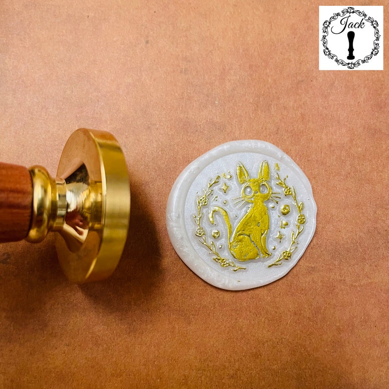 May include: A white wax seal with a gold design of a cat with a floral wreath around it. The seal is sitting on a brown surface.