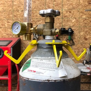 May include: A metal gas cylinder with a yellow bracket, pressure gauge, and flow meter. The cylinder has a brass valve and a silver handle. The gauge displays pressure readings. The background shows a workshop setting.