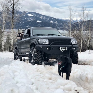 Può includere: Un pick-up nero con un paraurti nero e un cane nero in piedi di fronte ad esso. Il camion sta guidando su una strada innevata con montagne sullo sfondo.