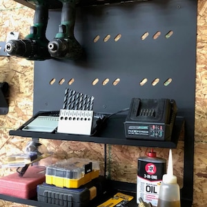 Puede incluir: Un estante de almacenamiento de metal negro montado en la pared con dos estantes. El estante superior contiene dos taladros inalámbricos verde y negro, un organizador de brocas y un cargador de batería negro y gris con el texto "MAX POWER FLEXPOWER". El estante inferior contiene una caja de herramientas roja, una caja de herramientas amarilla, una herramienta negra, una herramienta amarilla y negra, una botella de aceite 3 en 1 y una botella transparente con un líquido amarillo.