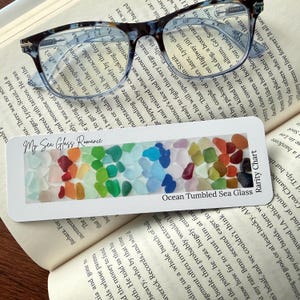 May include: A bookmark with the text "My Sea Glass Romance" and "Ocean Tumbled Sea Glass Rarity Chart" features a gradient of colorful sea glass pieces. The bookmark rests on an open book, with eyeglasses on top.