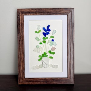 May include: A framed wall art piece featuring a bouquet of flowers crafted from various pieces of sea glass in shades of white, blue, green, and pink. The stems are drawn in thin gold lines, and the flowers are arranged in a white textured vase.