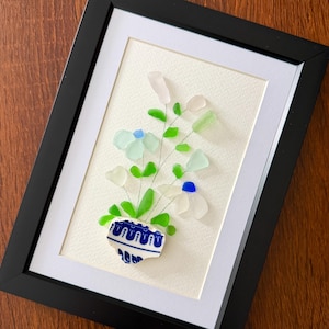 May include: Framed artwork featuring a floral arrangement crafted from sea glass. The flowers are white, light blue, and green, arranged in a blue and white ceramic vase. The artwork is set against a white background within a black frame.