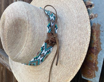 May include: A straw hat with a wide brim and a decorative beaded band in turquoise, white, and brown. The hat has a brown leather chin strap. The hat is hanging on a wooden fence.