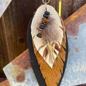 May include: A handcrafted leather pendant necklace in a leaf shape. The pendant features layers of black, tan, and cream-colored leather, with a feather and bead embellishment. The necklace has a brown leather cord.