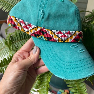 May include: Teal blue baseball cap with a colorful beaded band. The band features geometric patterns in red, white, yellow, and black.