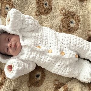 May include: A white crocheted baby onesie with a hood and ears. The onesie has wooden buttons and is lying on a brown and white patterned blanket.