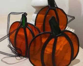 May include: Three stained glass pumpkins in orange and black with green stems. The pumpkins are hanging from black ribbon.