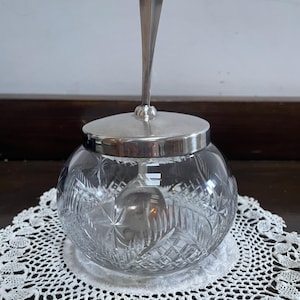 Könnte beinhalten: Ein klarer, runder Zuckerbehälter aus Glas mit einem silbernen Deckel und Löffel. Die Schüssel hat ein geschliffenes Glasdesign und steht auf einer weißen Spitzendecke. Der Löffel wird durch ein Loch im Deckel gesteckt.