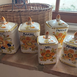 May include: A set of five square ceramic kitchen canisters with lids, featuring hand-painted orange floral patterns and blue accents. The canisters are labelled in French with the words CAFE, SUCRE, FARINE, SEL, and POIVRE in black script lettering.