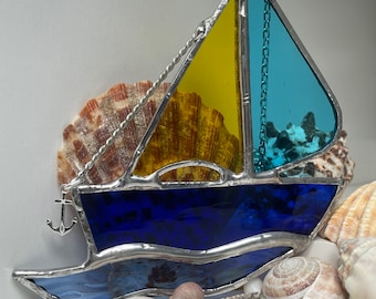 May include: A stained glass sailboat ornament with a blue hull, yellow sail, and turquoise sail. The ornament has a silver chain and anchor. The ornament is surrounded by seashells.