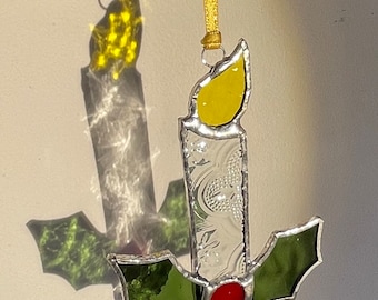 May include: A stained glass Christmas ornament shaped like a candle with a yellow flame and holly leaves. The candle is silver with a textured design and the holly leaves are green with a red berry.