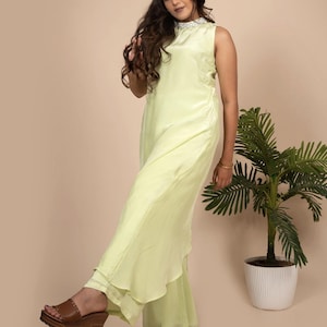 Könnte beinhalten: Ein hellgrünes, ärmelloses Maxikleid mit hohem Ausschnitt und einem weißen Perlenkragen. Das Kleid ist aus fließendem Stoff und hat einen mehrlagigen Rock. Das Model trägt braune Plateausandalen.