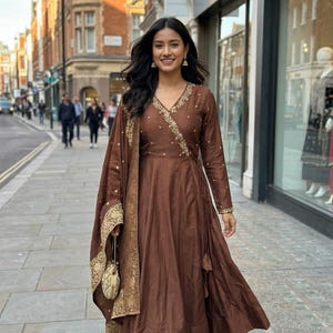 May include: A brown wrap dress with gold embroidery and a matching shawl. The dress has long sleeves and a flowing skirt. The shawl and dress are adorned with gold detailing. A small gold purse is held in the hand.