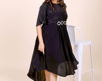 May include: A woman wearing a black dress with a high-low hemline and a silver belt. The dress has a pleated bodice and a flowing skirt. She is wearing clear heeled sandals.