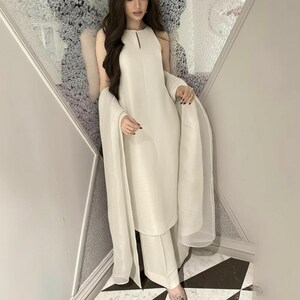 May include: A cream-colored, sleeveless dress and matching pants set with a sheer shawl. The dress has a keyhole neckline. The outfit is worn with sheer, patterned shoes. The background is a decorative wall and a black and white tiled floor.