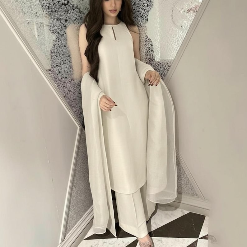 May include: A cream-colored, sleeveless dress and matching pants set with a sheer shawl. The dress has a keyhole neckline. The outfit is worn with sheer, patterned shoes. The background is a decorative wall and a black and white tiled floor.