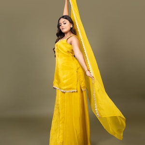 May include: A bright yellow sleeveless tunic and matching wide-leg trousers set. The tunic features a scoop neckline and a scalloped white trim along the hem. A long, sheer yellow dupatta scarf with a matching white scalloped border is held aloft.