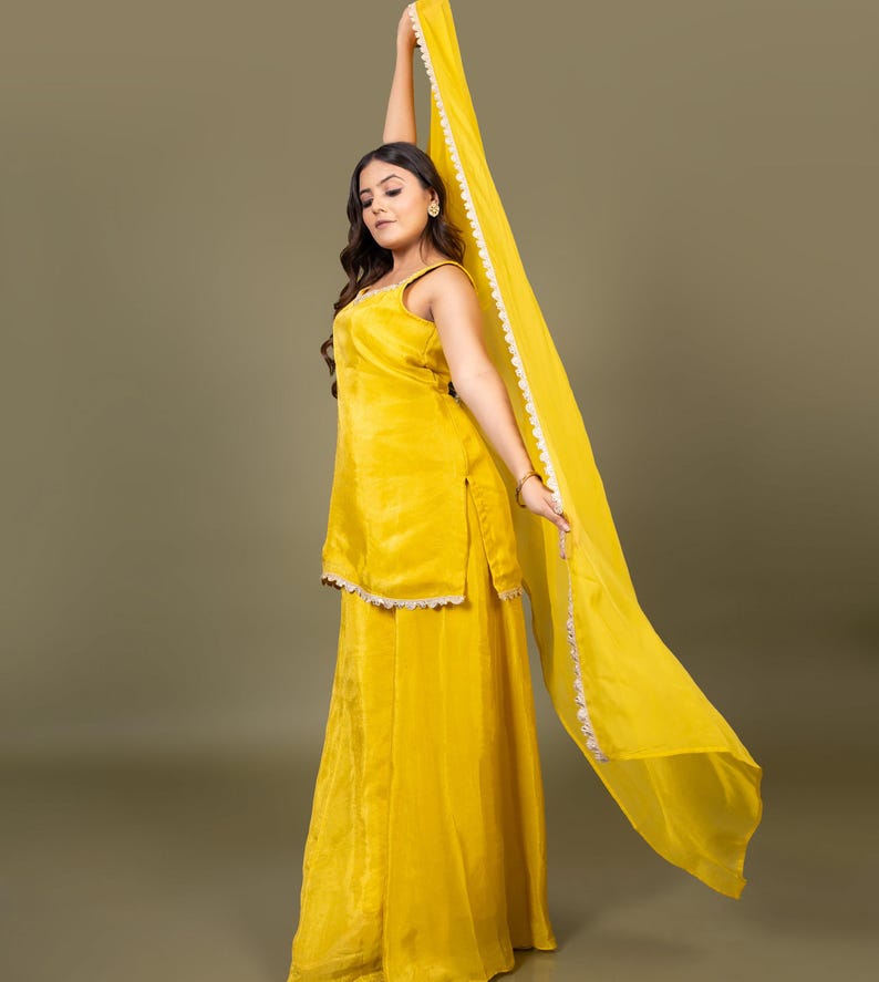 May include: A bright yellow sleeveless tunic and matching wide-leg trousers set. The tunic features a scoop neckline and a scalloped white trim along the hem. A long, sheer yellow dupatta scarf with a matching white scalloped border is held aloft.