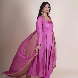 May include: A woman wearing a pink long-sleeved dress with gold embroidery and a matching pink sheer dupatta with gold trim. She is wearing gold sandals and has her hair pulled back.