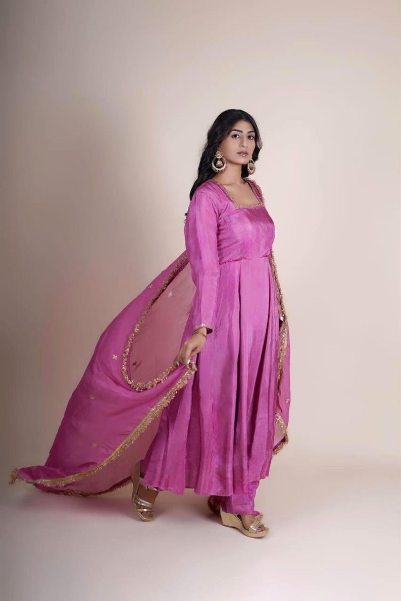 May include: A woman wearing a pink long-sleeved dress with gold embroidery and a matching pink sheer dupatta with gold trim. She is wearing gold sandals and has her hair pulled back.