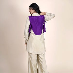 Op de afbeelding: Een vrouw draagt een cr&egrave;mekleurige top met een paarse vest en een bijpassende broek. Het vest heeft een unieke gebogen vorm en is aan de zijkanten vastgebonden.