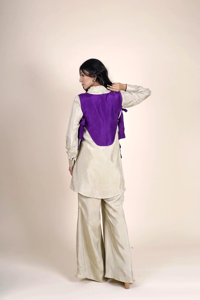 Op de afbeelding: Een vrouw draagt een cr&egrave;mekleurige top met een paarse vest en een bijpassende broek. Het vest heeft een unieke gebogen vorm en is aan de zijkanten vastgebonden.