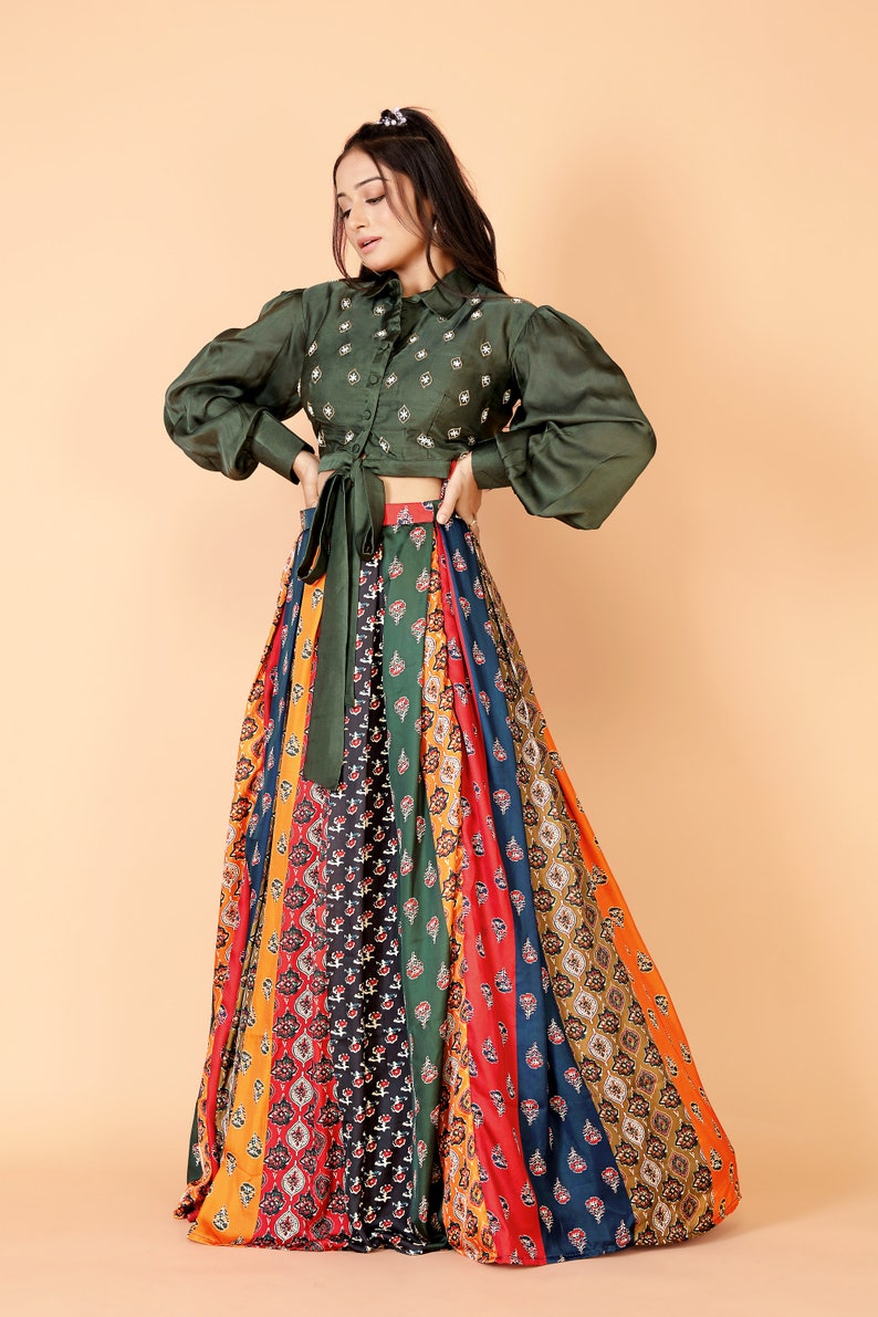 May include: An olive green, long-sleeved top with a collared neckline and embroidered details. The top is paired with a colorful, patterned maxi skirt featuring vertical stripes in various colors and designs. The outfit is a stylish ensemble.