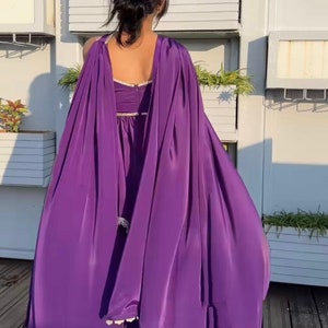 Puede incluir: Un vestido de sat&eacute;n morado con una larga sobrecapa tipo capa. El vestido tiene un ribete de encaje y un escote en V profundo.