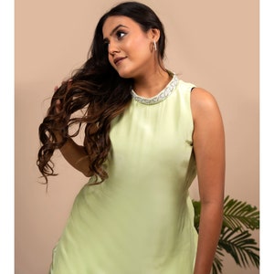 May include: A woman wearing a sleeveless, light green dress with a white beaded neckline. She has long, dark brown hair and is looking to the side.