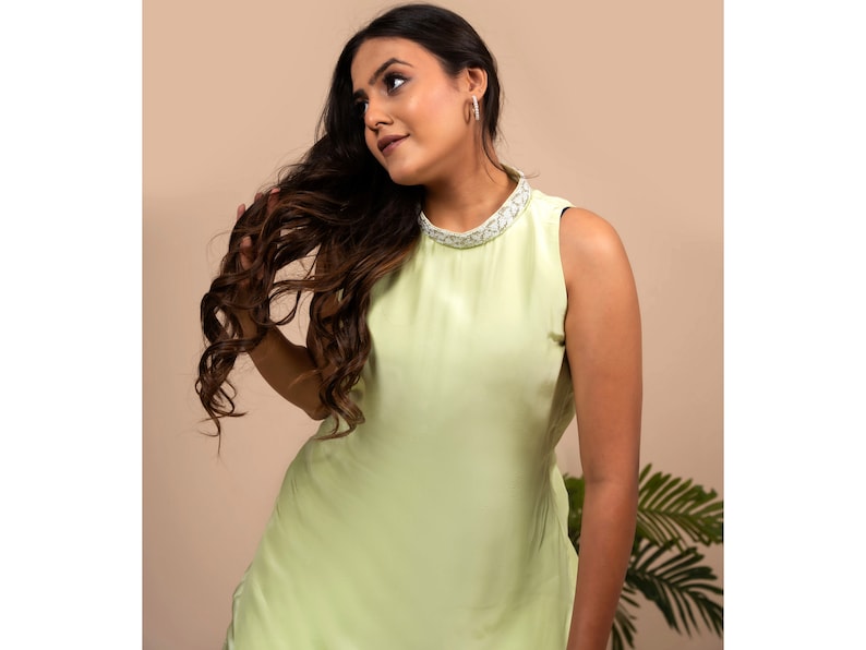 May include: A woman wearing a sleeveless, light green dress with a white beaded neckline. She has long, dark brown hair and is looking to the side.