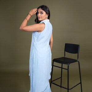 May include: A woman wearing a light blue sleeveless dress with a white collar and a white trim. She is standing in front of a black chair with a brown background.
