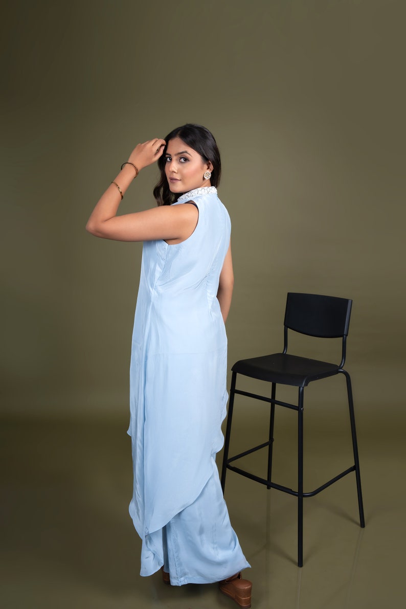 May include: A woman wearing a light blue sleeveless dress with a white collar and a white trim. She is standing in front of a black chair with a brown background.