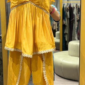 Puede incluir: Un conjunto de dos piezas de color amarillo mostaza con una blusa sin mangas y pantalones. La blusa tiene un escote de coraz&oacute;n y un dobladillo con volantes. Los pantalones tienen una pierna ancha y un ribete de encaje.