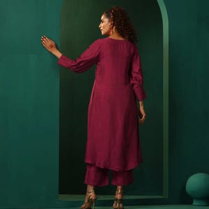 A maroon kurta and palazzo set. The kurta is a long-sleeved, knee-length top with a relaxed fit. The palazzo pants are loose-fitting and ankle-length. The outfit is made from a soft, flowing fabric. The model is wearing gold heels.