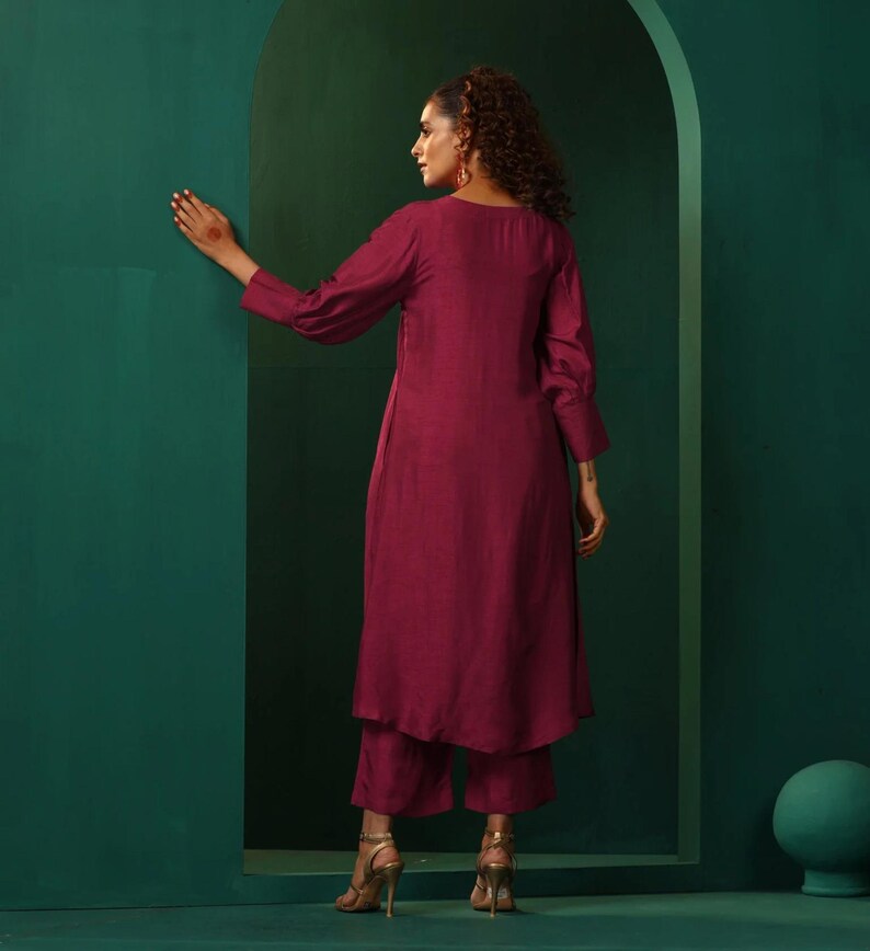 A maroon kurta and palazzo set. The kurta is a long-sleeved, knee-length top with a relaxed fit. The palazzo pants are loose-fitting and ankle-length. The outfit is made from a soft, flowing fabric. The model is wearing gold heels.