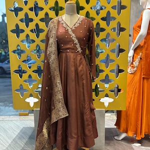May include: A brown wrap dress with long sleeves and a V-neck, adorned with delicate white embroidery along the neckline. A matching shawl with gold detailing drapes over the dress. The dress is displayed on a mannequin.