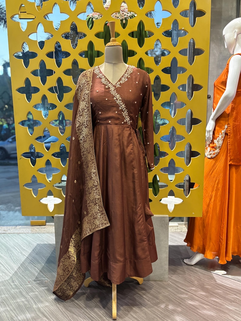 May include: A brown wrap dress with long sleeves and a V-neck, adorned with delicate white embroidery along the neckline. A matching shawl with gold detailing drapes over the dress. The dress is displayed on a mannequin.