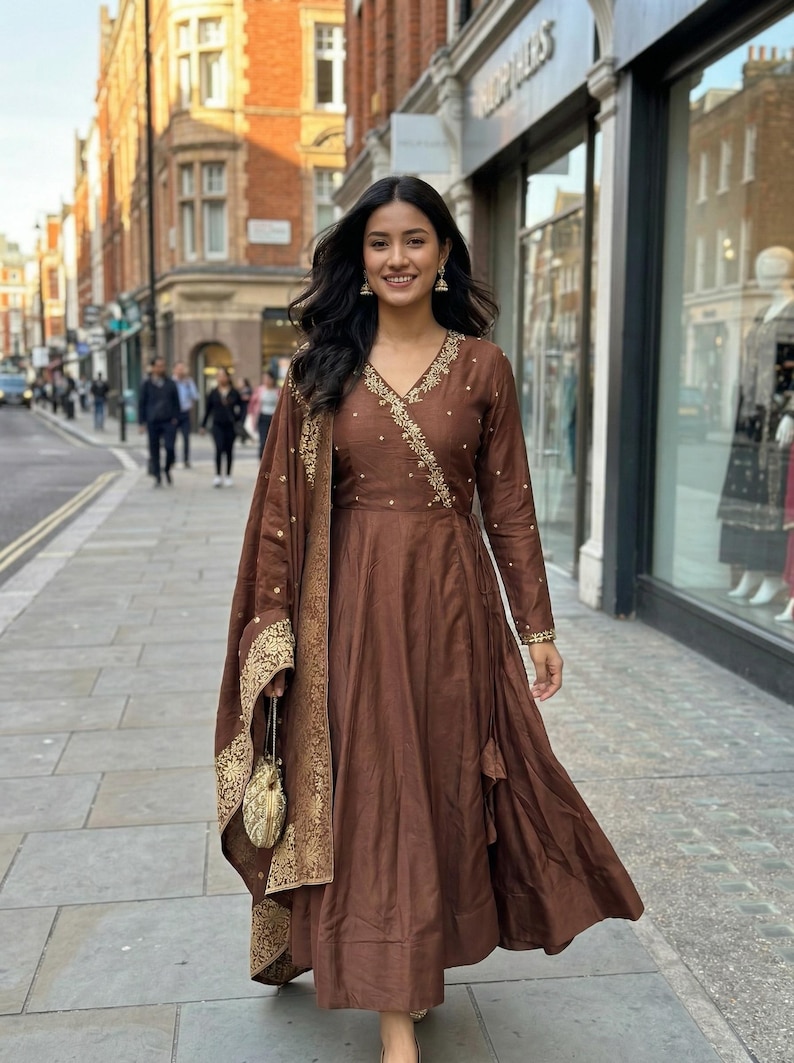 May include: A brown wrap dress with gold embroidery and a matching shawl. The dress has long sleeves and a flowing skirt. The shawl and dress are adorned with gold detailing. A small gold purse is held in the hand.