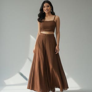 May include: A brown two-piece outfit featuring a crop top with spaghetti straps and high-waisted, wide-leg pants. The outfit is made of a flowing fabric and is suitable for a variety of occasions. The model is wearing gold sandals.