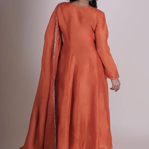 May include: An orange-colored, flowing dress with long sleeves and a matching scarf. The dress has a simple design with a slight sheen, and the scarf is trimmed with silver detailing. The outfit is a traditional style.