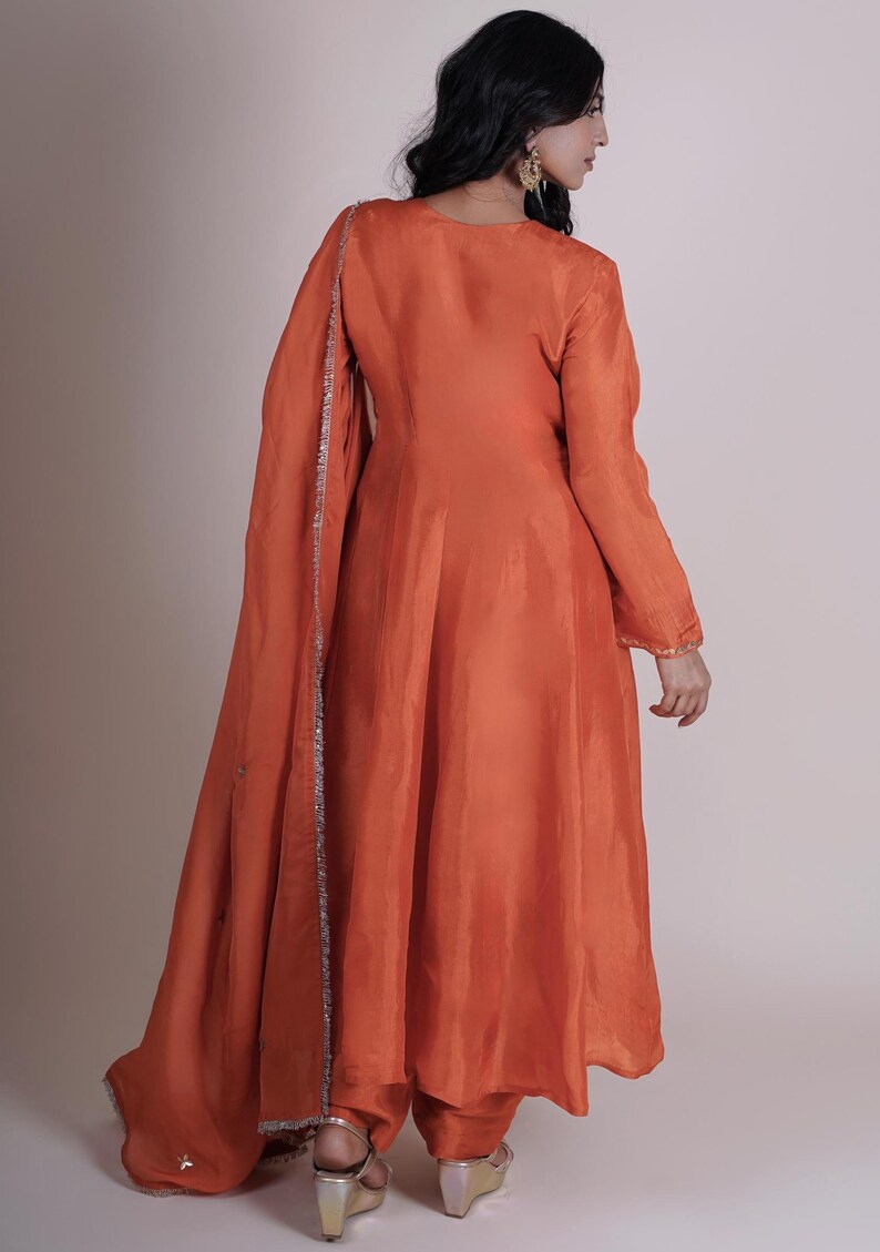 May include: An orange-colored, flowing dress with long sleeves and a matching scarf. The dress has a simple design with a slight sheen, and the scarf is trimmed with silver detailing. The outfit is a traditional style.