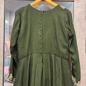 Pu&ograve; includere: Abito verde oliva con maniche lunghe e scollo tondo. L'abito presenta ricami dorati sui polsini e sulle spalle. L'abito &egrave; appeso a un'appendiabiti di legno.