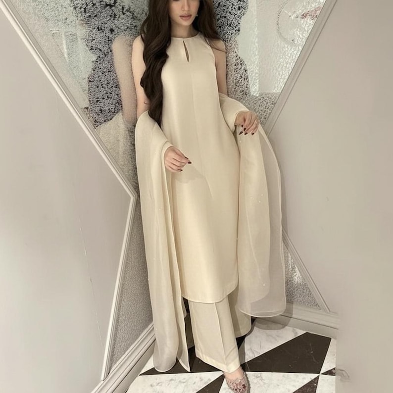 May include: A cream-colored, sleeveless outfit consisting of a tunic and wide-leg pants. The tunic has a keyhole neckline. A sheer shawl drapes over the shoulders. The outfit is worn with embellished shoes.