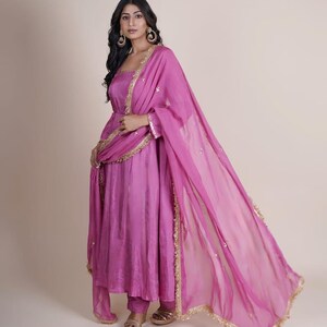 May include: A woman wearing a pink long-sleeved dress with gold trim and a matching pink sheer dupatta. The dress has a gathered skirt and the dupatta has a gold fringe.
