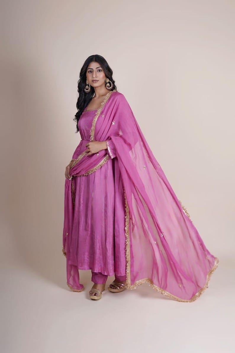 May include: A woman wearing a pink long-sleeved dress with gold trim and a matching pink sheer dupatta. The dress has a gathered skirt and the dupatta has a gold fringe.