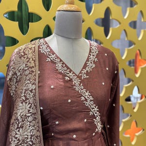 May include: A brown wrap dress with long sleeves and a V-neck, adorned with white embroidery along the neckline. The dress features small, scattered white embellishments and is paired with a matching shawl with a gold floral pattern.