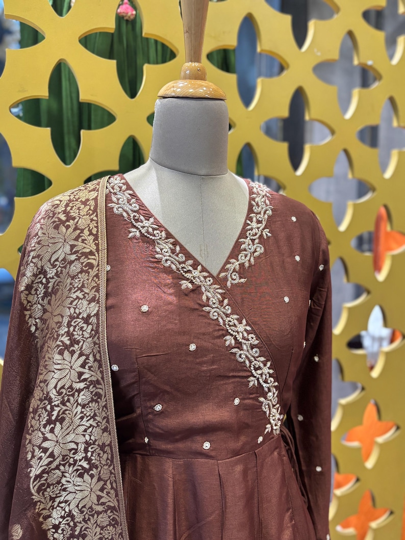 May include: A brown wrap dress with long sleeves and a V-neck, adorned with white embroidery along the neckline. The dress features small, scattered white embellishments and is paired with a matching shawl with a gold floral pattern.