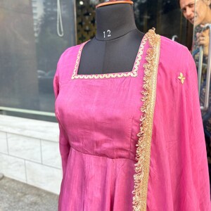 May include: A pink dress with gold trim and embroidery. The dress has a square neckline and a flowing skirt. The dress is displayed on a mannequin.