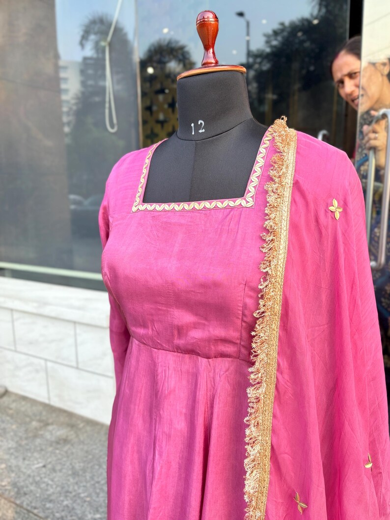 May include: A pink dress with gold trim and embroidery. The dress has a square neckline and a flowing skirt. The dress is displayed on a mannequin.